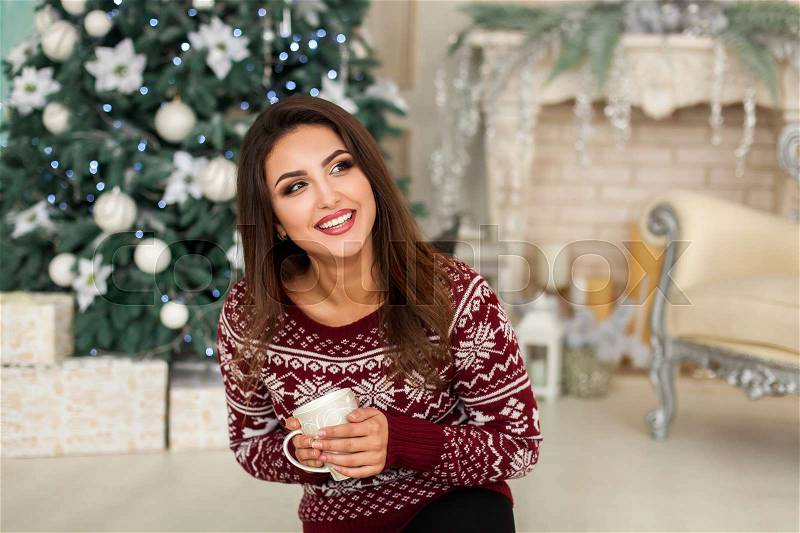 Model sitting with cup of drink near christmas tree in living room, stock photo