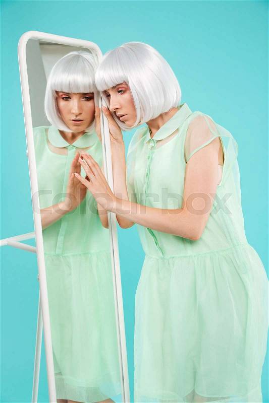 Beautiful young woman in blonde wig standing and touching mirror, stock photo