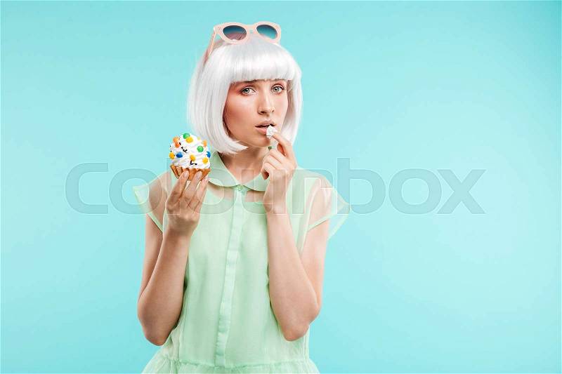 Beautiful young woman in blonde wig standing and eating cupcake over blue background, stock photo