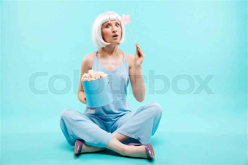 Amazed young woman with marshmallows and magic stick sitting with legs crossed, stock photo
