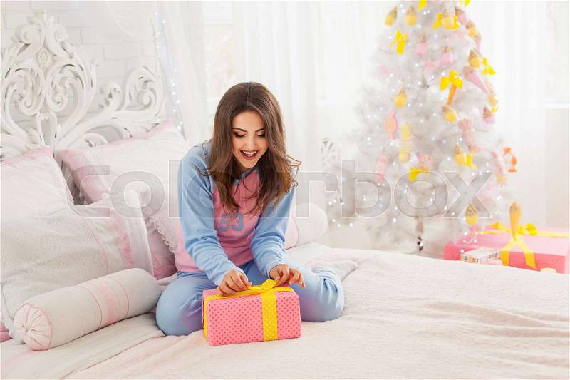 Laughing model sitting on the bed and opening christmas present, stock photo