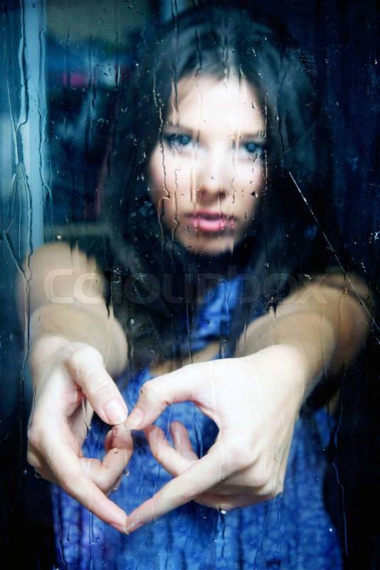 Young girl making heart from fingers behind wet window, stock photo