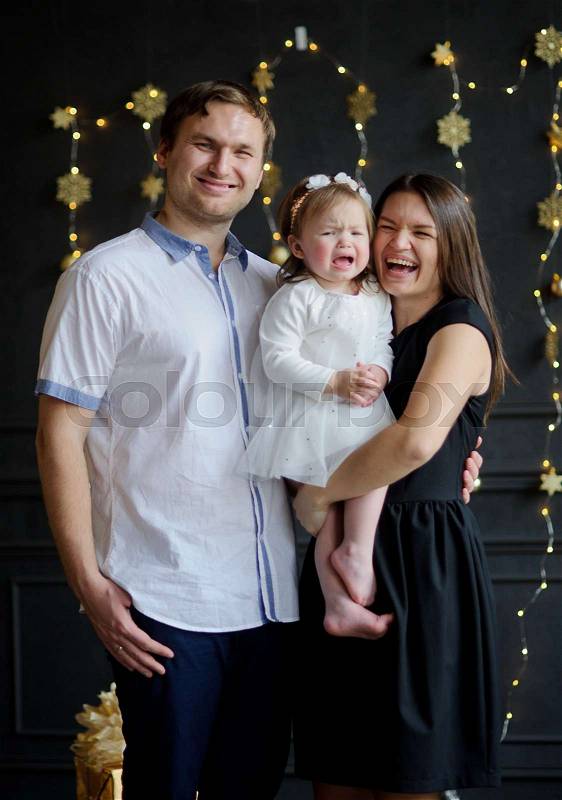 Young parents calm the crying child. The room is festively decorated. Little girl cries on hands at mother. Baby is dressed in a white dress, on the head of her a wreath. Parents look in the camera and smile, stock photo