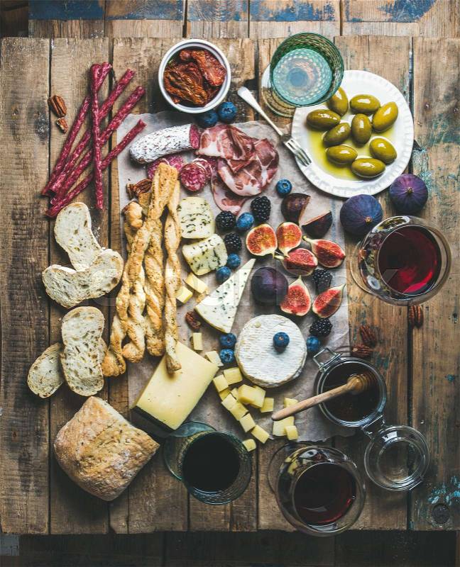 Wine and snack set with various wines in glasses, meat variety, bread, sun-dried tomatoes, honey, green olives, figs, nuts and fresh berries on wax paper over rustic wooden table background, top view, stock photo