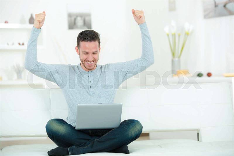 Cheerful young man with laptop raising hands indoors, stock photo