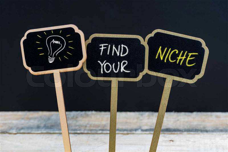 Concept message FIND YOUR NICHE and light bulb as symbol for idea written with chalk on wooden mini blackboard labels, defocused chalkboard and wood table in background, stock photo