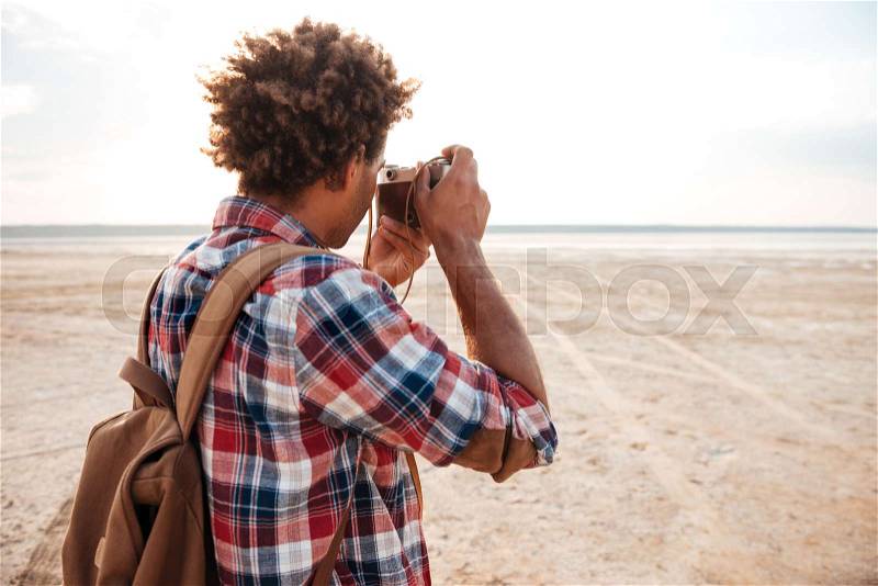 Back view of african american young man with backpack taking photos on the beach, stock photo
