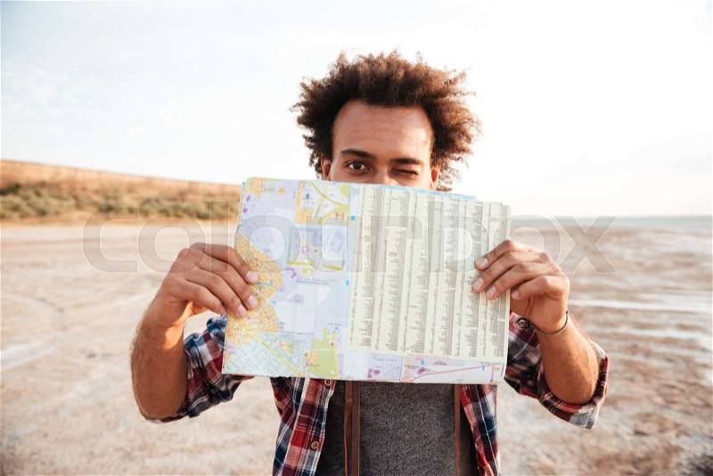 Joyful african american young man covered his face with map and winking, stock photo