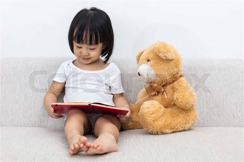 Asian Chinese little girl reading book with teddy bear on the sofa in the living room, stock photo