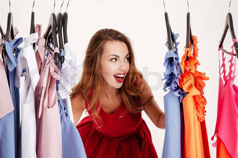 Beautiful smiling blonde woman standing inside wardrobe rack full of clothes looking away, stock photo