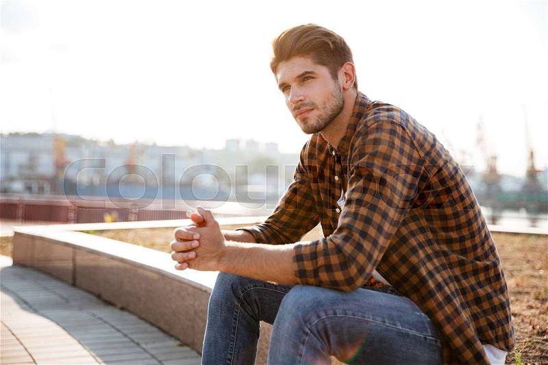 Pensive young man in plaid shirt sitting and thinking outdoors, stock photo