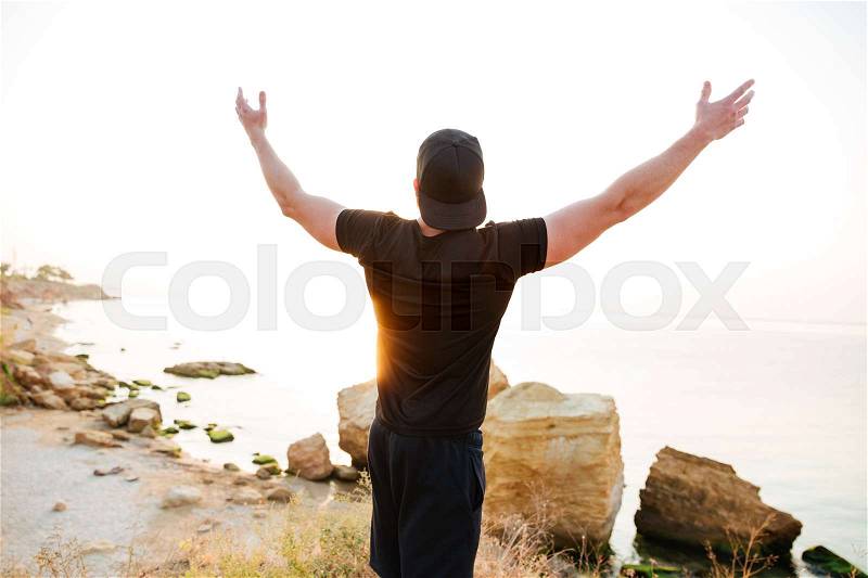 Back view of a young man standing with arms raised at the rocky beach by the sea looking at sunset, stock photo