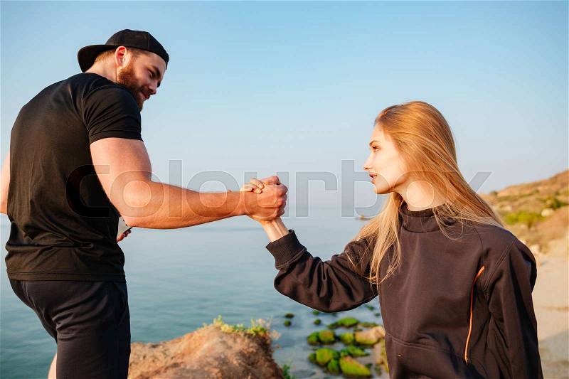 Healthy bearded man helping his girlfriend to climb a hill at the seaside, stock photo