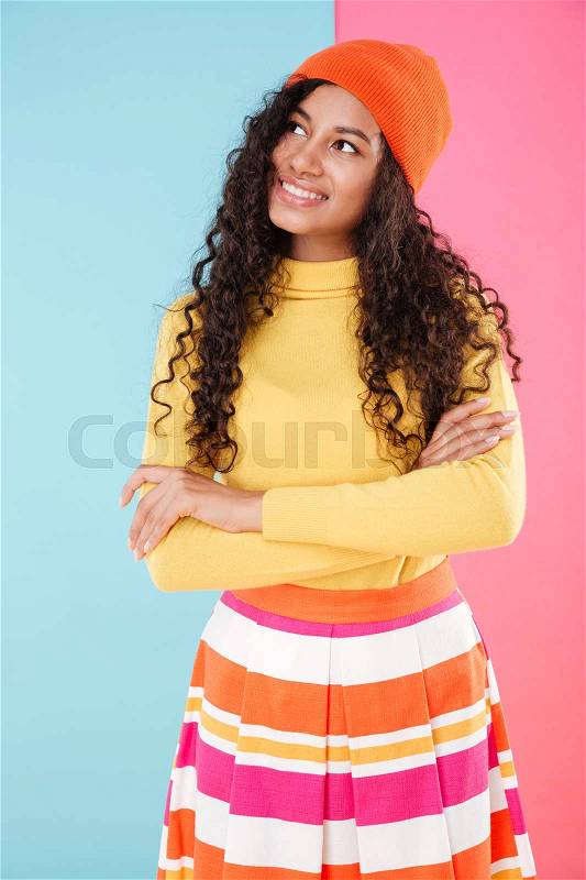 Cute curly african young woman in hat standing with arms crossed and thinking over colorful background, stock photo