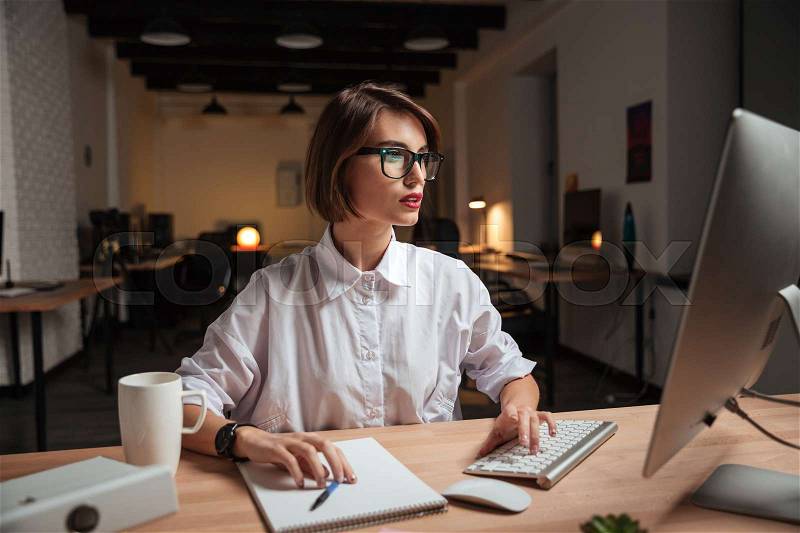 Concentrated attractive young businesswoman in glasses working with computer in office, stock photo