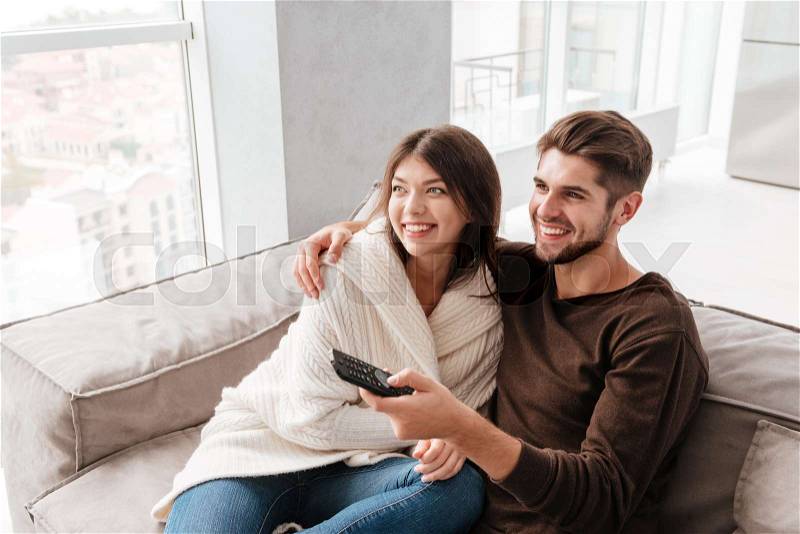 Cheerful cute young couple hugging and watching TV on couch at home, stock photo