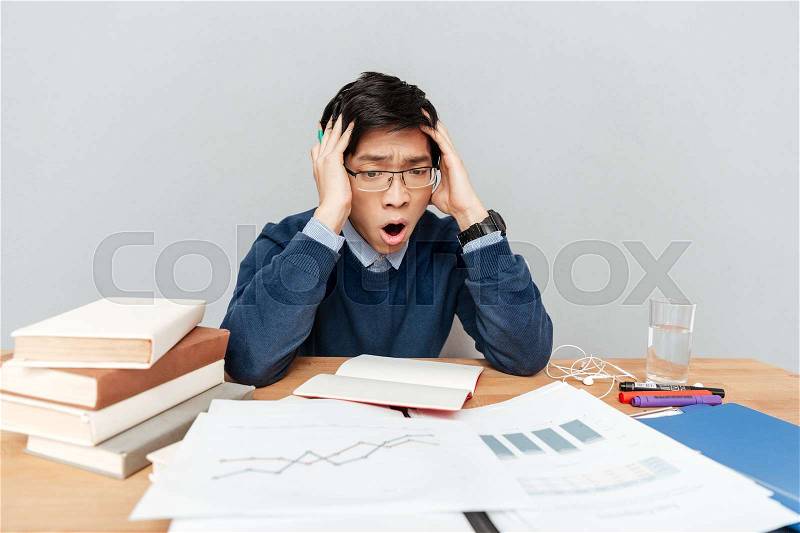 Asian student in panic with notebook. in glasses. gray background. sits by the table, stock photo