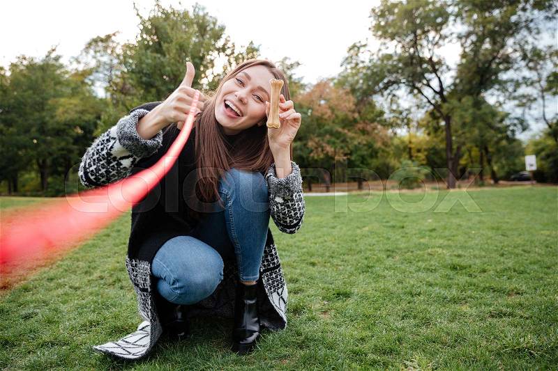 Smiling attractive young woman holding bone for her dog and showing thumbs up in park, stock photo