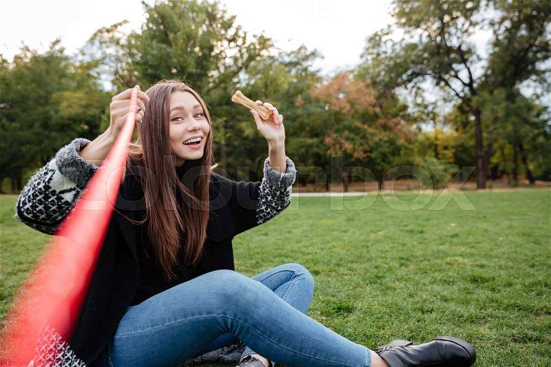 Smiling relaxed young woman sitting and throwing bone to her dog in park, stock photo