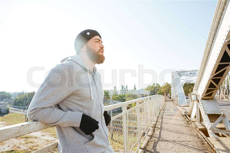 Tired young man athlete in hat resting after jogging outdoors, stock photo