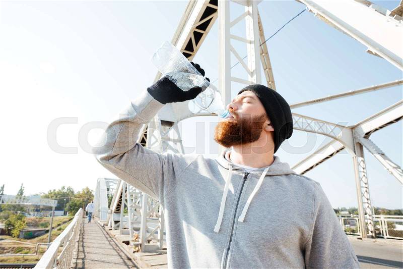 Portrait of a young bearded athlete in sportswear drinking water while standing at the bridge, stock photo