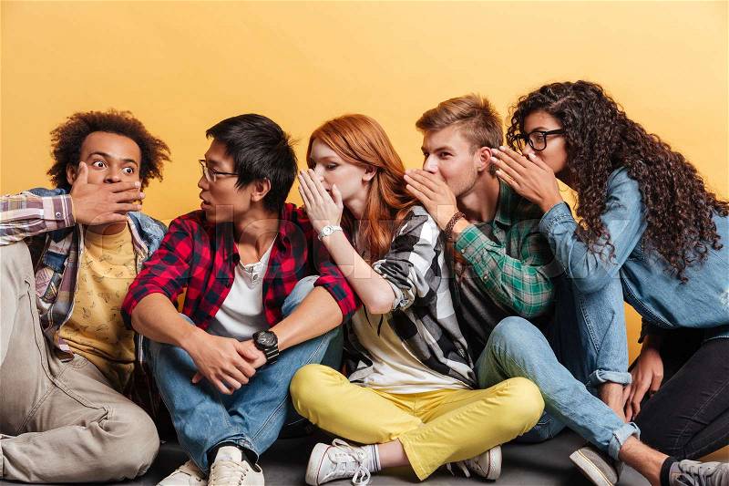 Multiethnic group of young people sitting and telling secrets to each other over yellow background, stock photo
