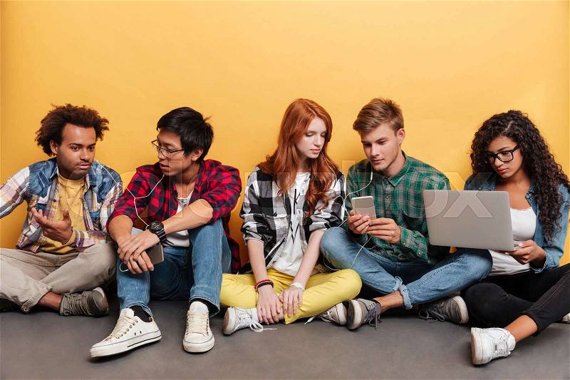 Multiethnic group of serious young friends listening to music from cell phone and using laptop over yellow background, stock photo