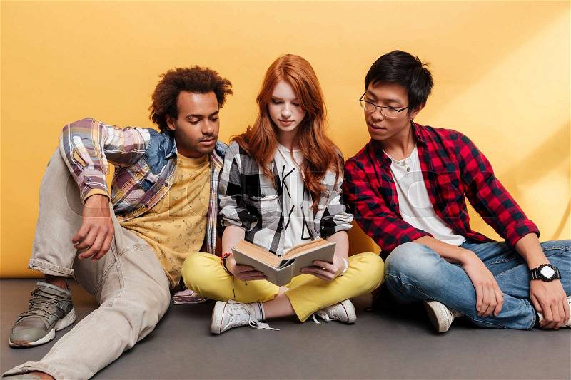 Three serious young people sitting and reading book together over yellow background, stock photo