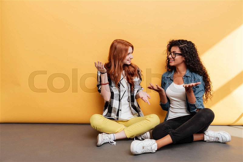 Two smiling confused young women sitting with legs crossed and talking over yellow background, stock photo