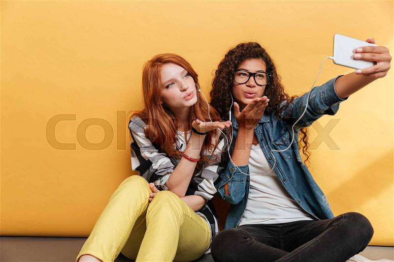 Two cute young women sending kiss and making selfie with mobile phone over yellow background, stock photo