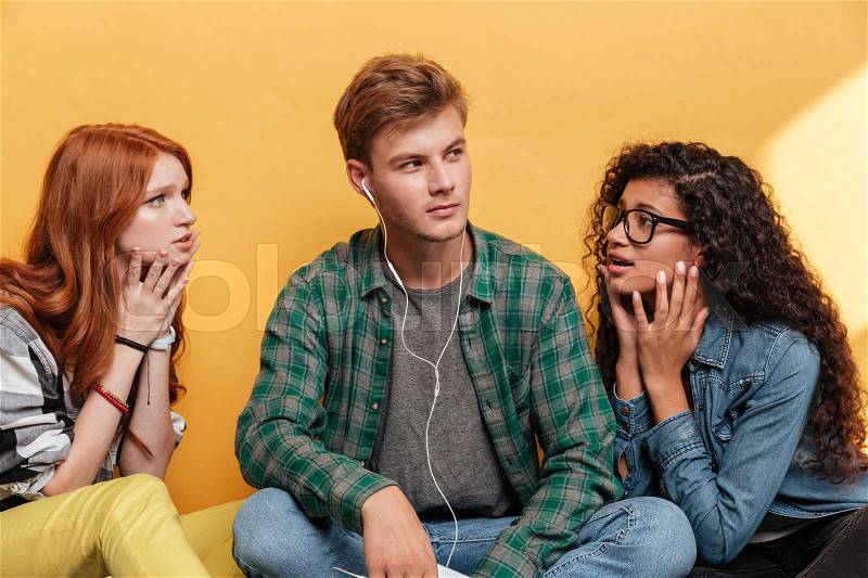 Love triangle of two young women in love and attractive man listening to music over yellow background, stock photo