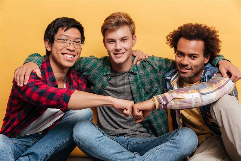 Three happy young men friends sitting and hugging over yellow background, stock photo