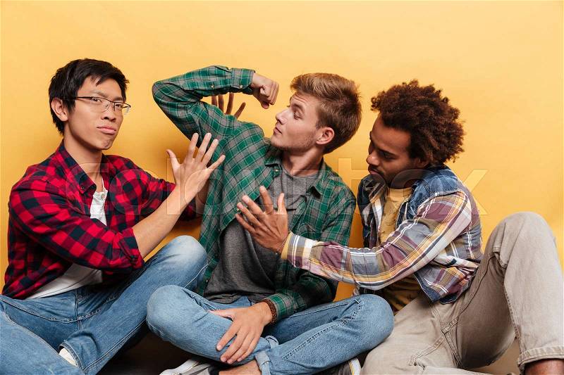 Three funny young men friends showing biceps and having fun over yellow background, stock photo