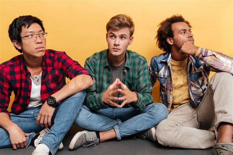 Three thoughtful handsome young men friends sitting and thinking over yellow background, stock photo