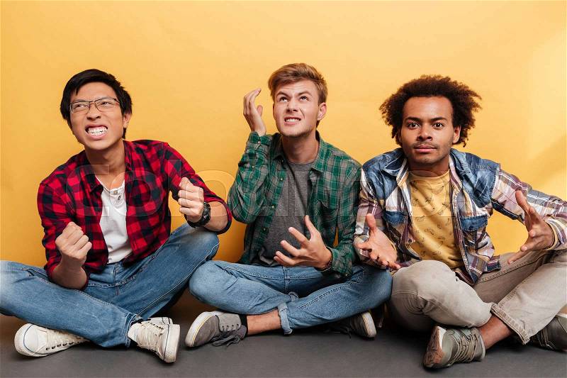 Three angry irritated young men friends sitting with legs crossed over yellow background, stock photo