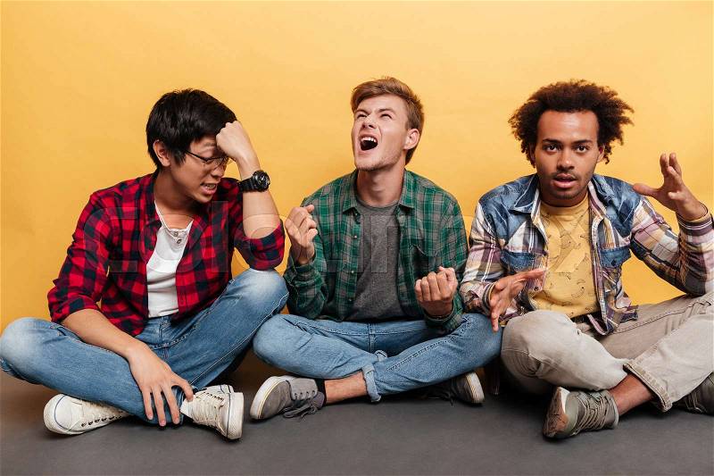 Three mad furious young men friends sitting and shouting over yellow background, stock photo