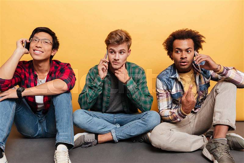 Three handsome young men friends sitting and talking on cell phone over yellow background, stock photo