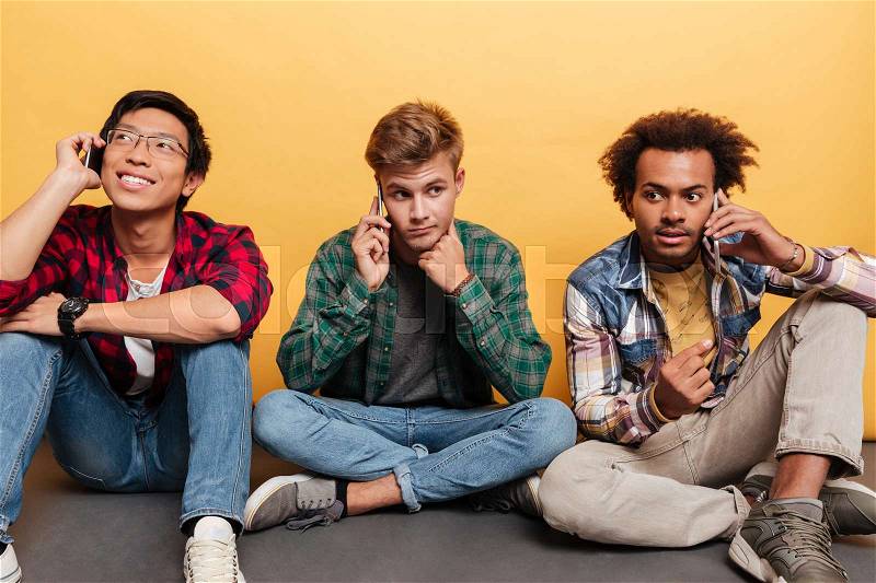 Multiethnic group of three attractive young men friends sitting and talking on mobile phone over yellow background, stock photo