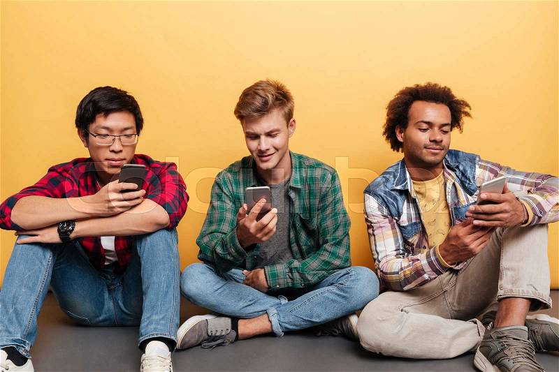 Multiethnic group of three young men friends sitting and using mobile phone over yellow background, stock photo