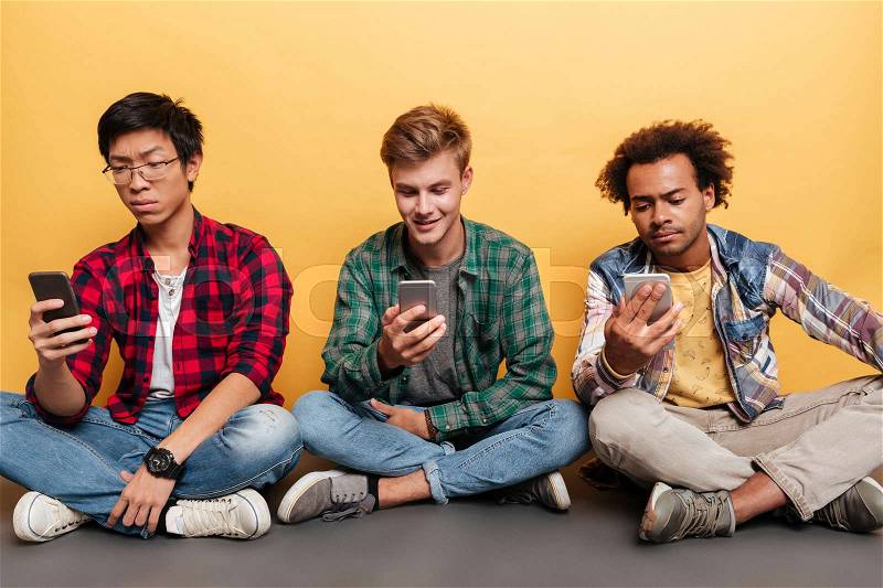 Three handsome young men friends using cell phone over yellow background, stock photo