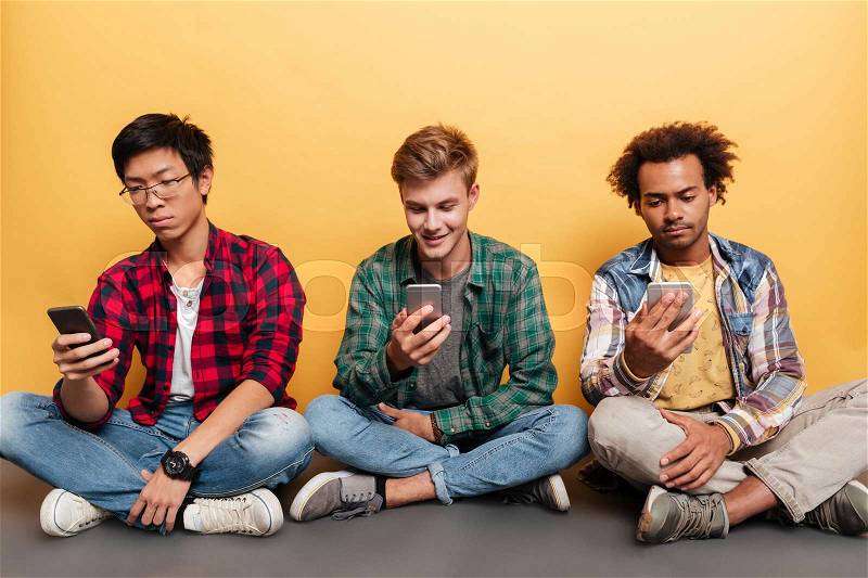 Three young men friends sitting and using smartphone over yellow background, stock photo