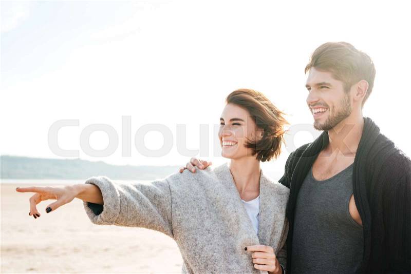 Laughing casual couple standing at the beach and pointing finger away, stock photo