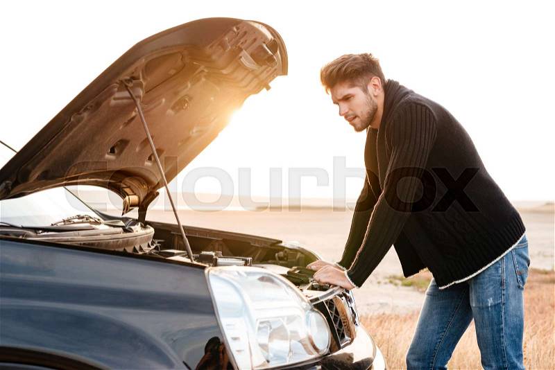 Young upset casual man trying to fix his broken car outdoors, stock photo