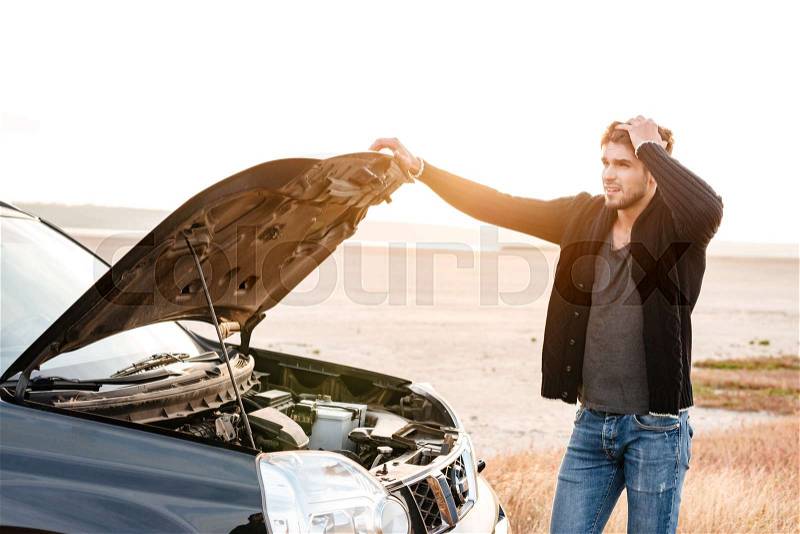 Concentrated young man holding hands on vehicle hood and looking inside it while standing outdoors, stock photo