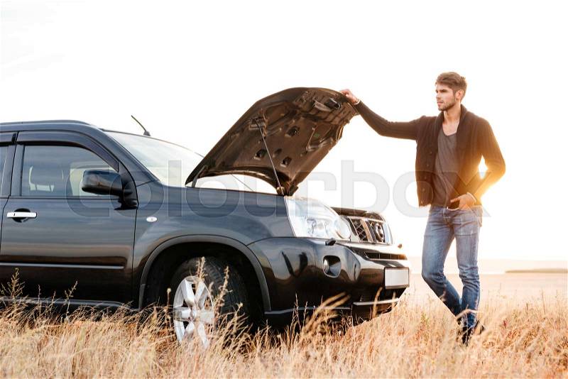 Concentrated young man holding hands on vehicle hood and looking inside it while standing outdoors, stock photo