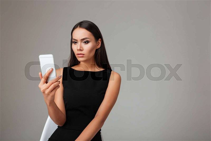 Portrait of a young brunette woman in black dress looking at mobile phone over gray background, stock photo