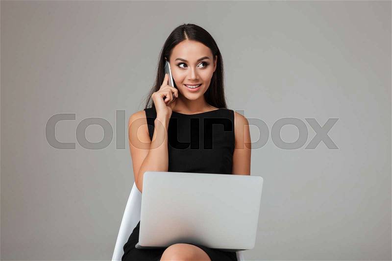 Happy young businesswoman sitting on the chair with laptop and talking on mobile phone over gray background, stock photo