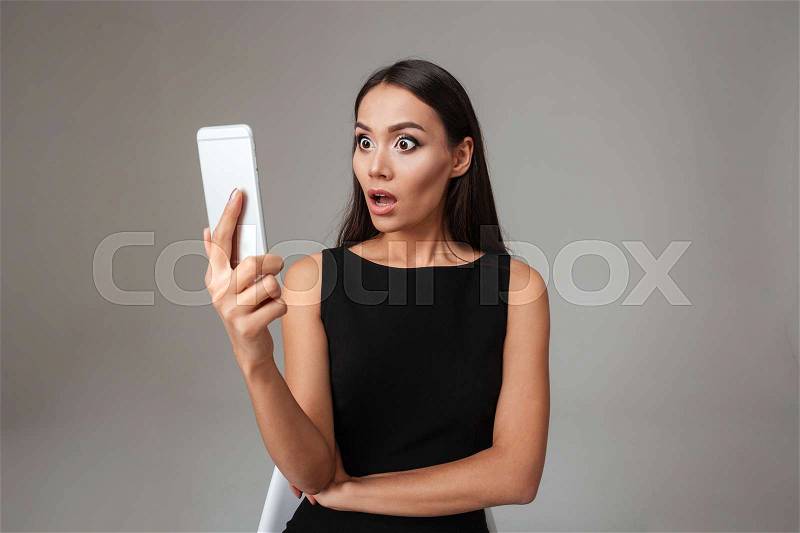 Shocked young woman in black dress looking at mobile phone over gray background, stock photo