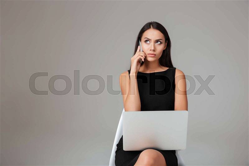 Displeased young brunette woman sitting with laptop and talking on mobile phone over gray background, stock photo