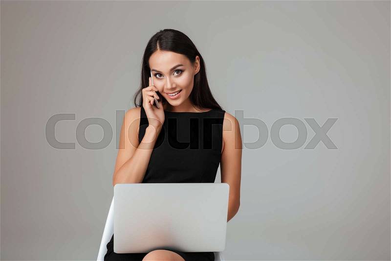 Happy young businesswoman sitting on the chair with laptop and talking on mobile phone over gray background, stock photo